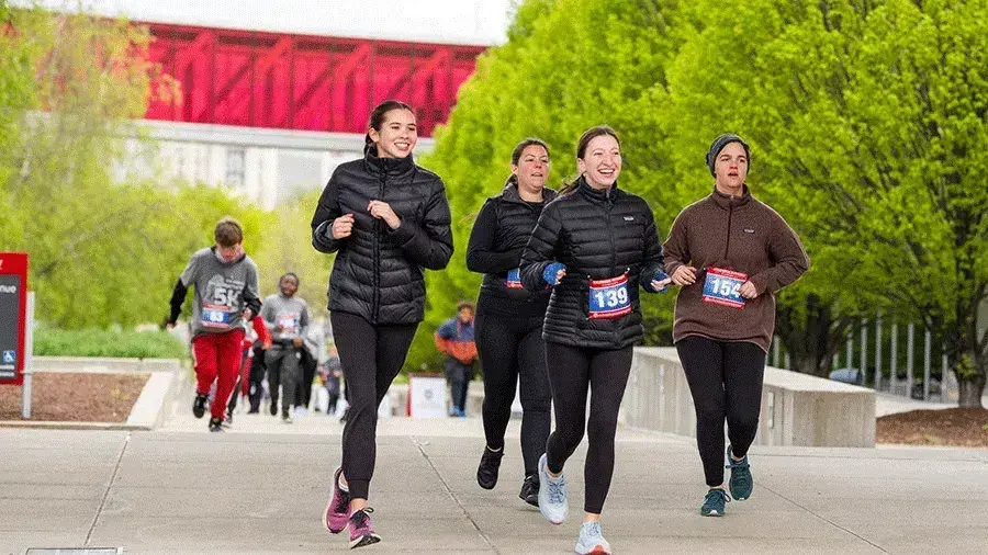 Participants run along a paved path during an organized race, with several runners wearing numbered race bibs and surrounded by green trees and a red building in the background.