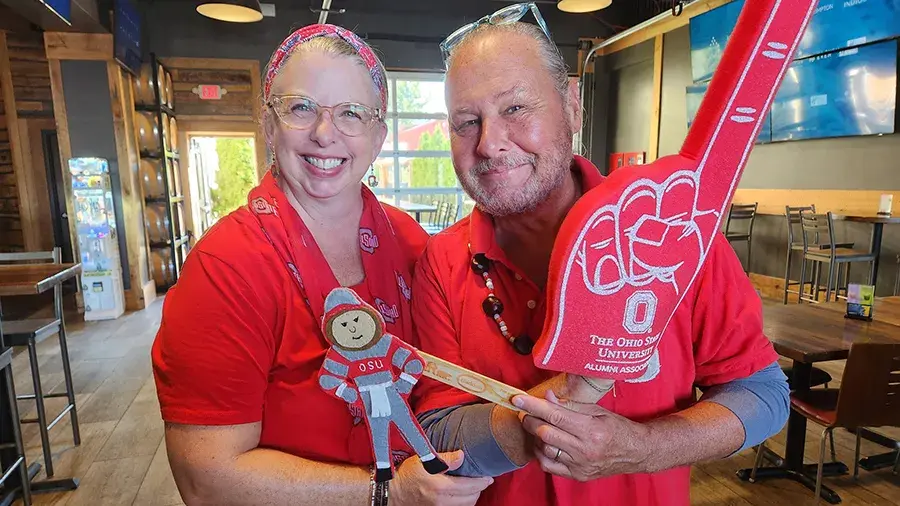 Two people in red Ohio State–themed shirts stand inside a restaurant, holding a Brutus Buckeye cutout and a large foam finger with “The Ohio State University Alumni Association” printed on it.