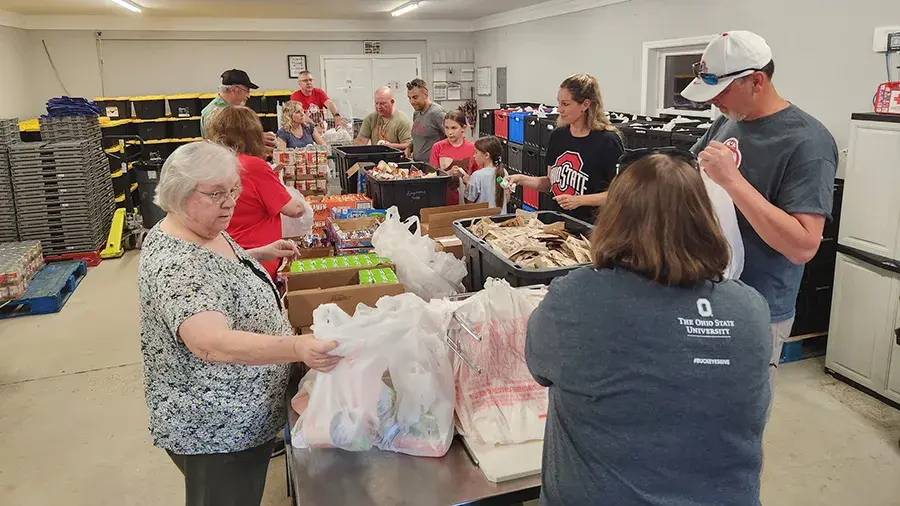 A group of people work together in a community space, organizing and packing food items such as canned goods, fresh produce, and bagged groceries on long tables.
