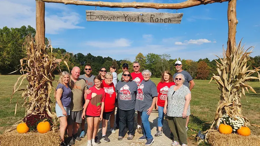 A group of people stands under a wooden archway decorated with dried cornstalks and pumpkins. The sign above the arch reads “Empower Youth Ranch.” The group is gathered outdoors in a grassy field with trees in the background on a sunny day.