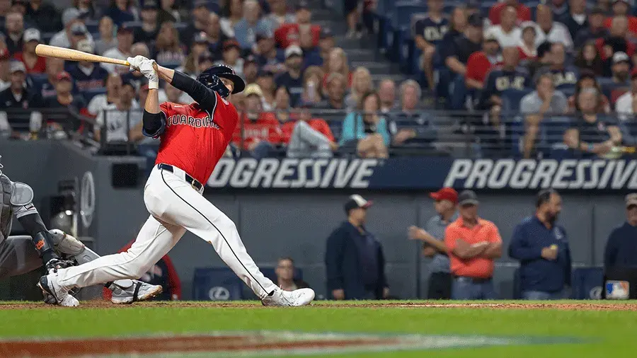 A Guardians baseball player in a red jersey swings at a pitch during a game, with a crowded stadium in the background.