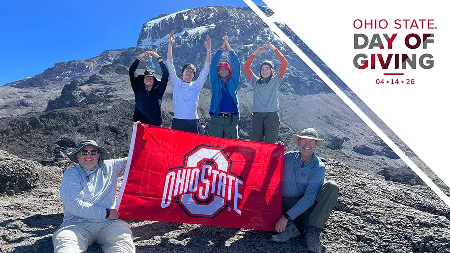 A group of hikers on a rocky mountain slope hold an Ohio State flag while some stand behind them forming the O‑H‑I‑O pose. A graphic on the right reads Ohio State Day of Giving 04‑14‑26.