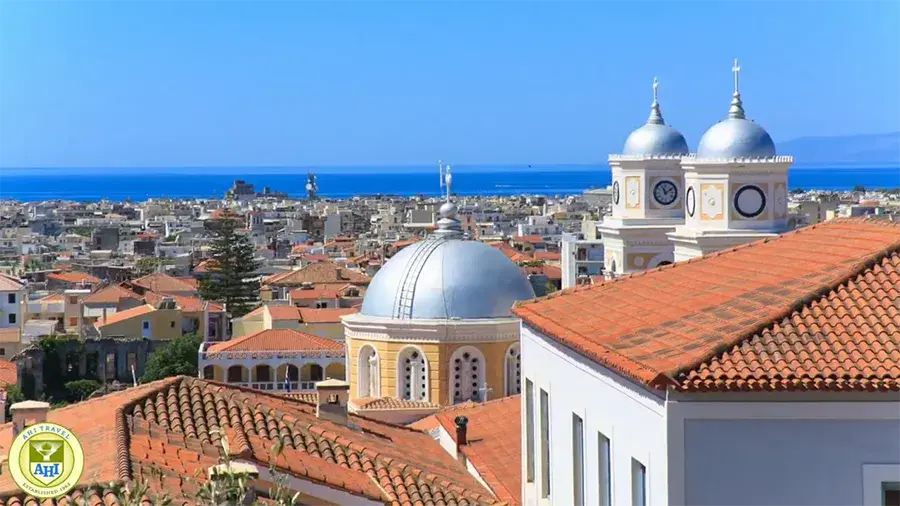 A coastal city with red-tiled rooftops, domed church towers, and the blue sea in the background under a clear sky.