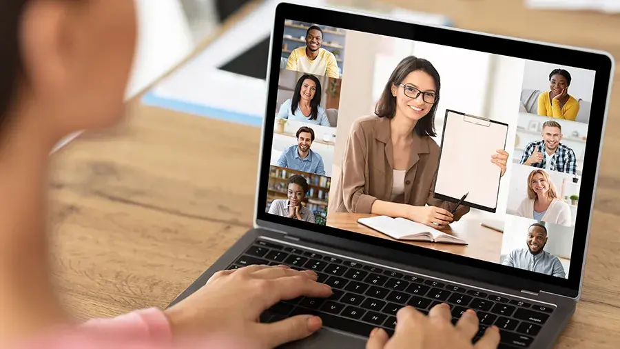 A person sitting at a desk uses a laptop showing a video call with multiple participants in a grid layout. The person on the laptop screen is holding up a clipboard or notepad during the virtual meeting.