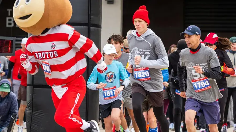 A group of runners participating in a race, including Brutus Buckeye. The runners wear race bibs and athletic clothing as they move forward together.
