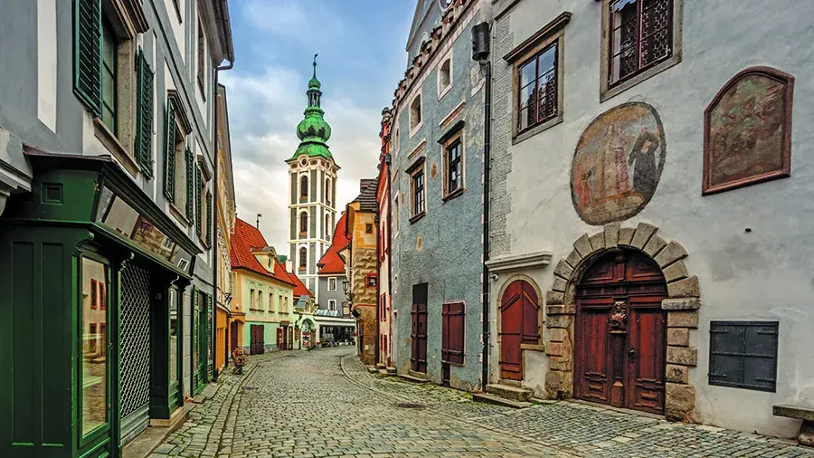 A narrow cobblestone street lined with old European buildings, leading toward a tall church tower with a green spire.