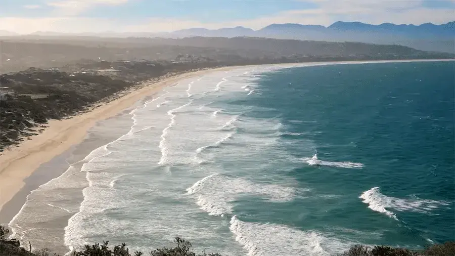 A wide sandy beach with gentle waves rolling in from a blue ocean, with mountains and a coastal town in the distance.