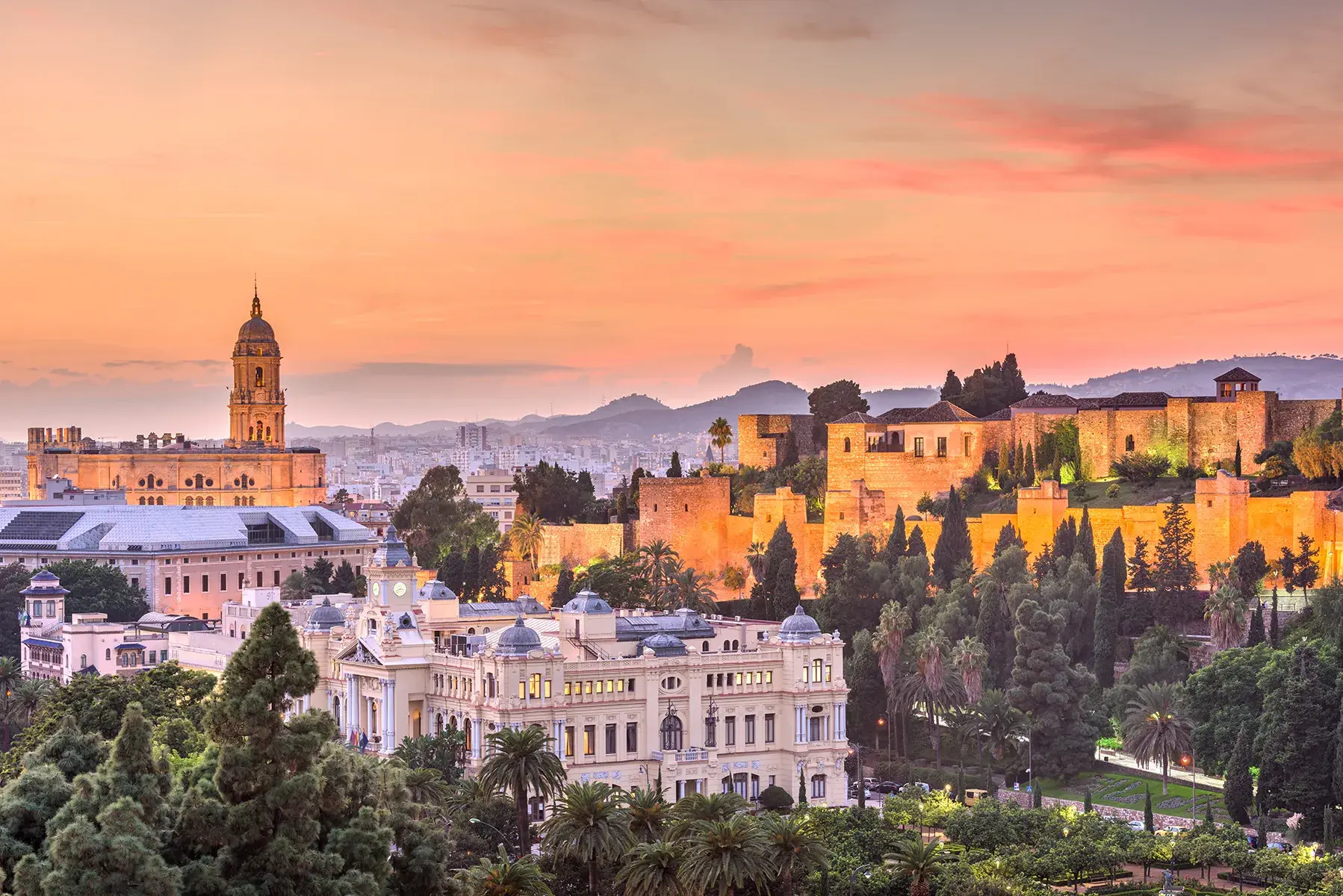Sunset view of a historic cityscape with illuminated buildings, palm trees, and hills in the background.
