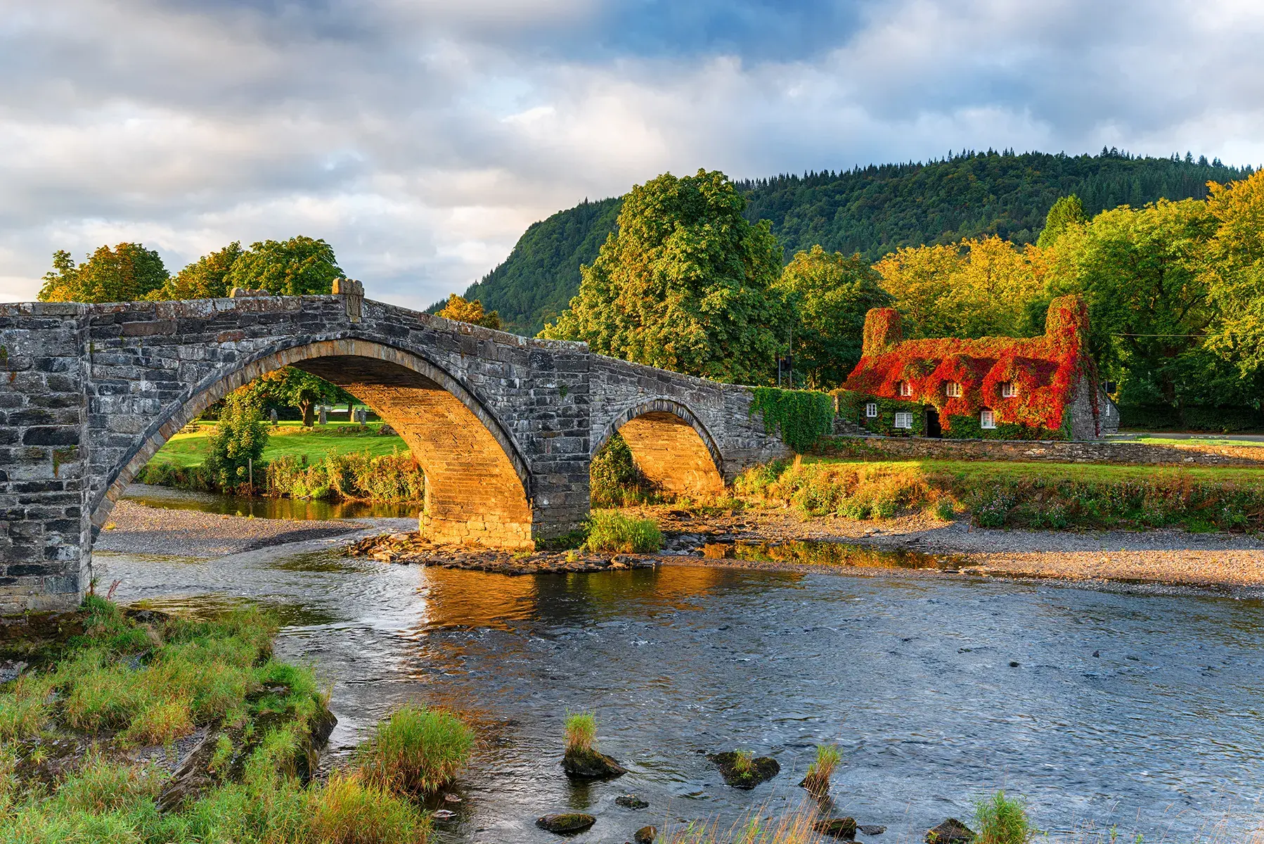 Stone bridge over a shallow river with a small ivy-covered cottage and trees in the background at sunset.
