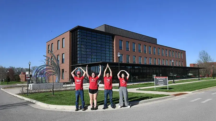 Four people in red shirts stand on a lawn in front of a modern brick campus building, posing with their arms to spell O‑H‑I‑O.