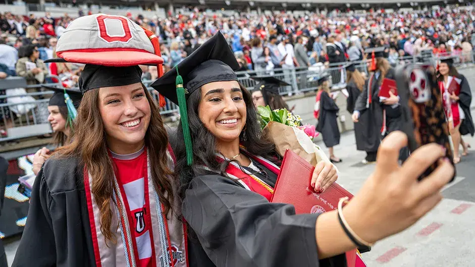 Two people in graduation caps and gowns take a selfie on a football stadium field, with one holding a diploma and bouquet and the other wearing Brutus’ giant cap on top of her graduation cap. A crowd of graduates and guests fills the stands in the background.