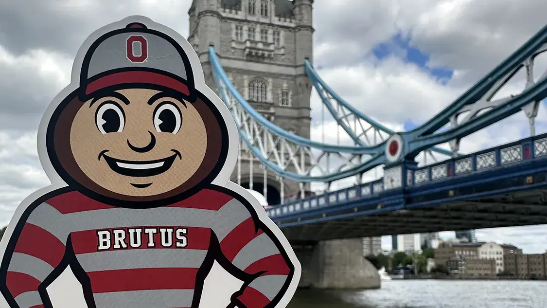 A hand holds a Brutus Buckeye sticker in a red‑and‑gray Ohio State uniform in front of London’s Tower Bridge over the River Thames on a cloudy day.
