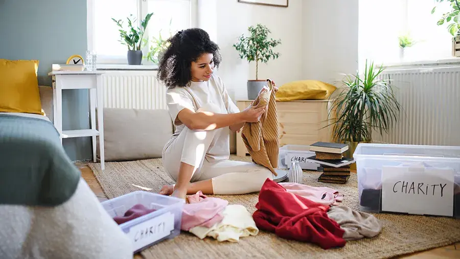 A person sits on the floor of a bright living room, sorting clothes into clear bins labeled “charity,” with folded clothing and boxes around them.