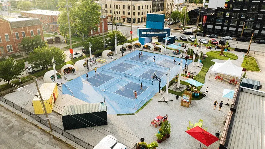 Aerial view of Gravity Experience Park with several blue sports courts, people playing games, seating areas with small tents, and shipping containers, surrounded by city streets and buildings.