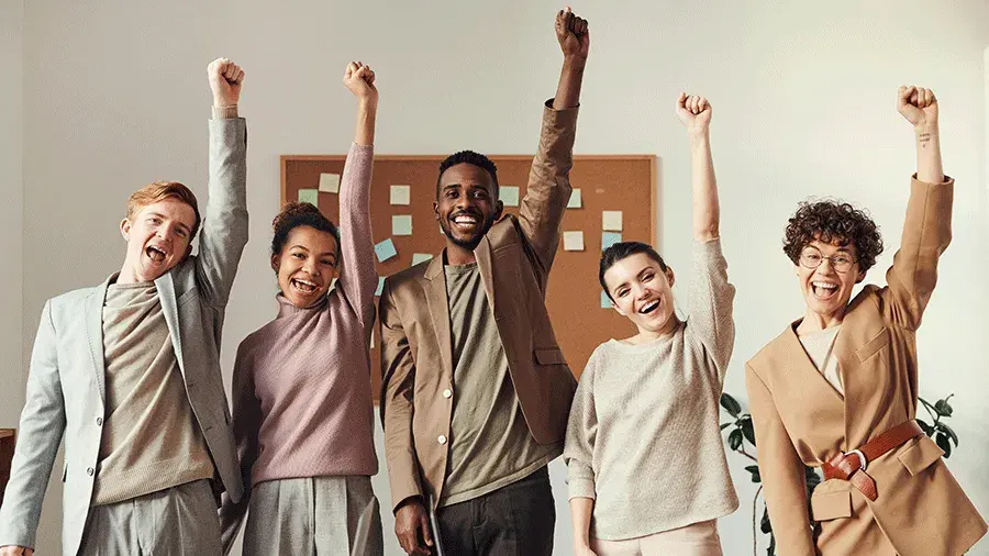 Five people stand side by side in an office setting with arms raised and fists up. A corkboard with sticky notes and a plant are visible in the background.