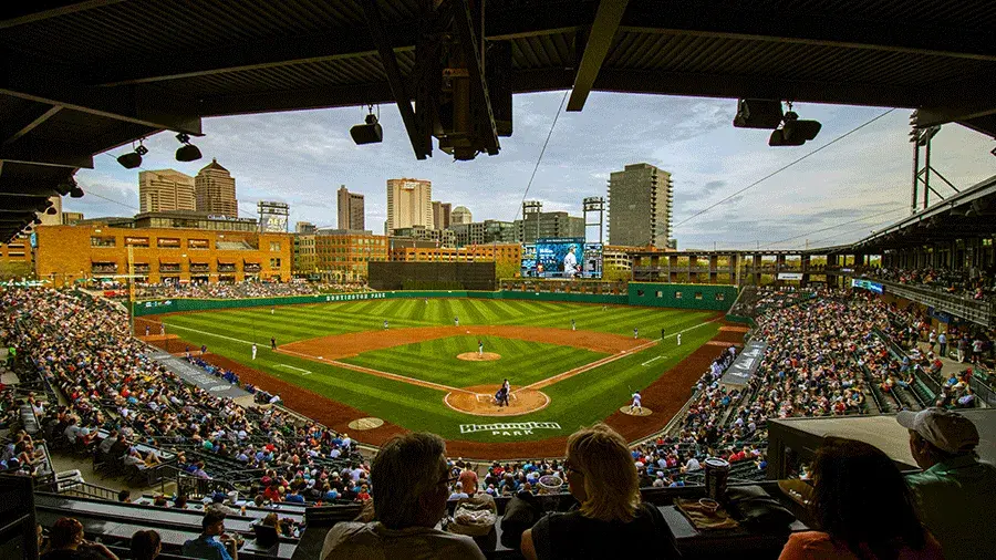 Wide view of a baseball game at Huntington Park, showing the green field, players on the diamond, packed seating on both sides, and the Columbus skyline in the background beneath a stadium roof.