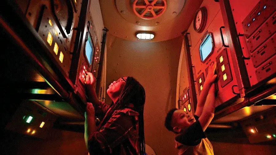 Two kids playing inside a narrow submarine, red-lit control room, reaching up to press glowing buttons and screens on tall panels filled with lights and switches.
