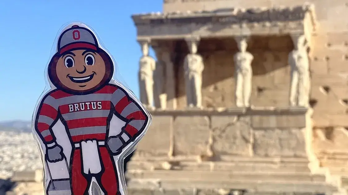 A hand holds a Brutus Buckeye sticker wearing a red‑and‑gray Ohio State uniform, with ancient stone ruins and carved statues in the background under a blue sky.