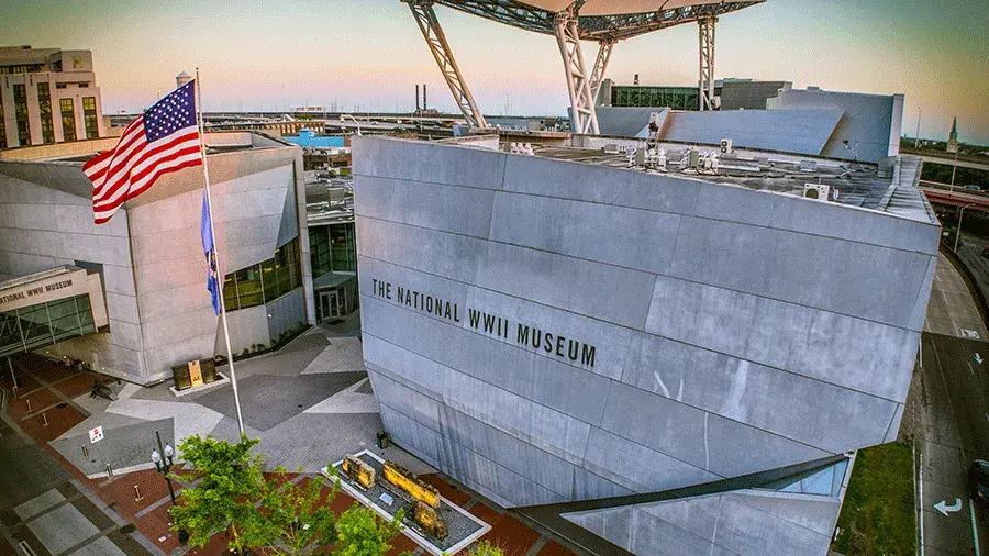Exterior of The National WWII Museum with large gray buildings, an American flag on a flagpole and the museum name visible on the wall.
