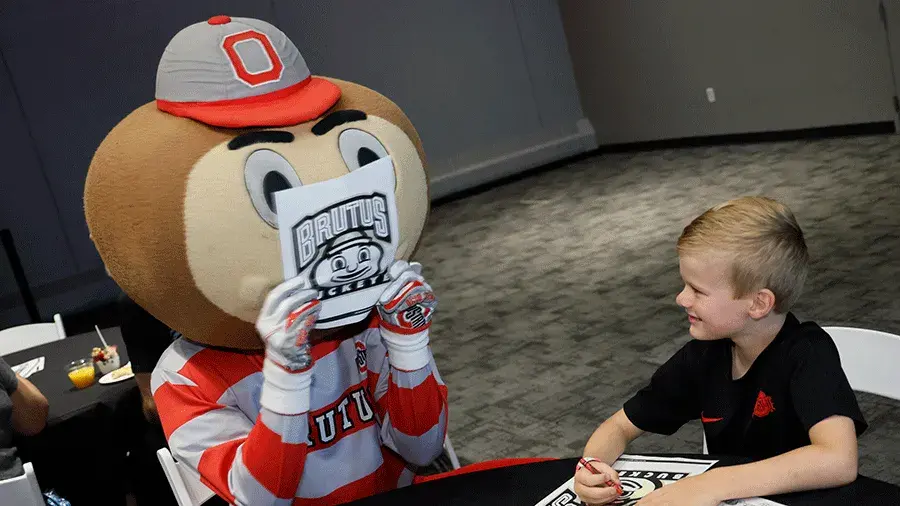 Brutus Buckeye holding a “Brutus Buckeye” coloring page and sitting with a child at a table at an indoor event.