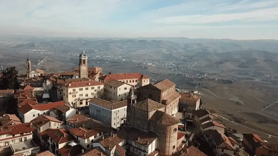 Aerial view of a historic hilltop town with red-tiled roofs and a church tower, overlooking rolling countryside and distant hills.