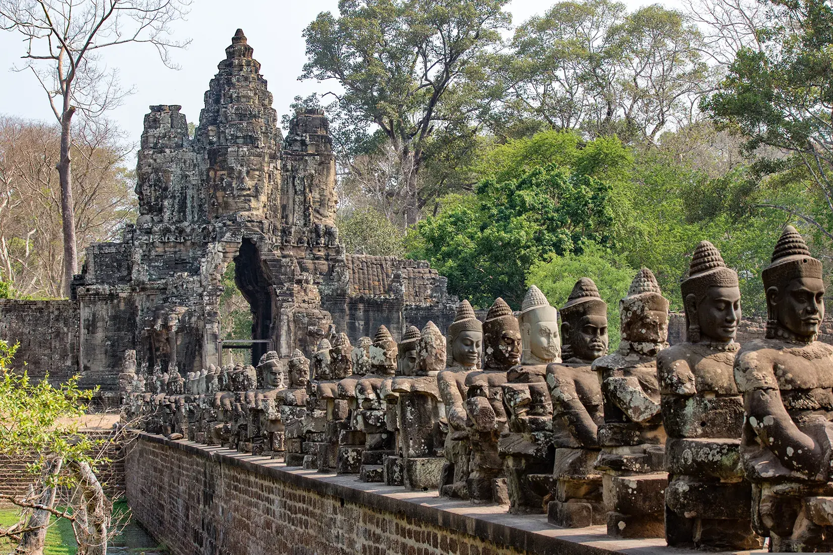 Stone statues line a long bridge leading to an ancient stone temple gate, with weathered carvings, mossy surfaces, and tall trees surrounding the ruins.