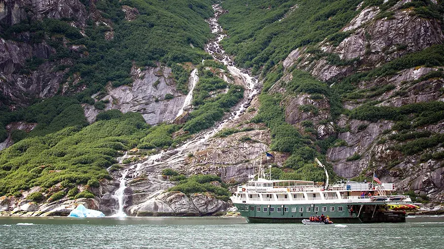 A white tour boat floats on calm water near a steep, green mountainside with a tall waterfall cascading down rocky cliffs into the sea.