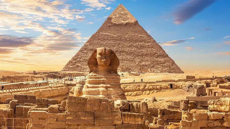 Stone Sphinx in the foreground with a large pyramid behind it, surrounded by desert ruins under a blue sky.
