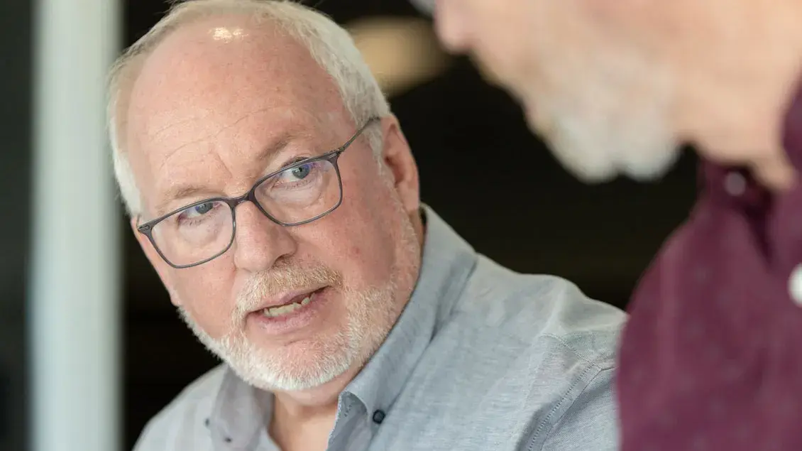 Dale Gelter talks to a colleague with a serious expression. He’s an older man with neat white hair and glasses and wears a button-down shirt with an embroidered Block O.