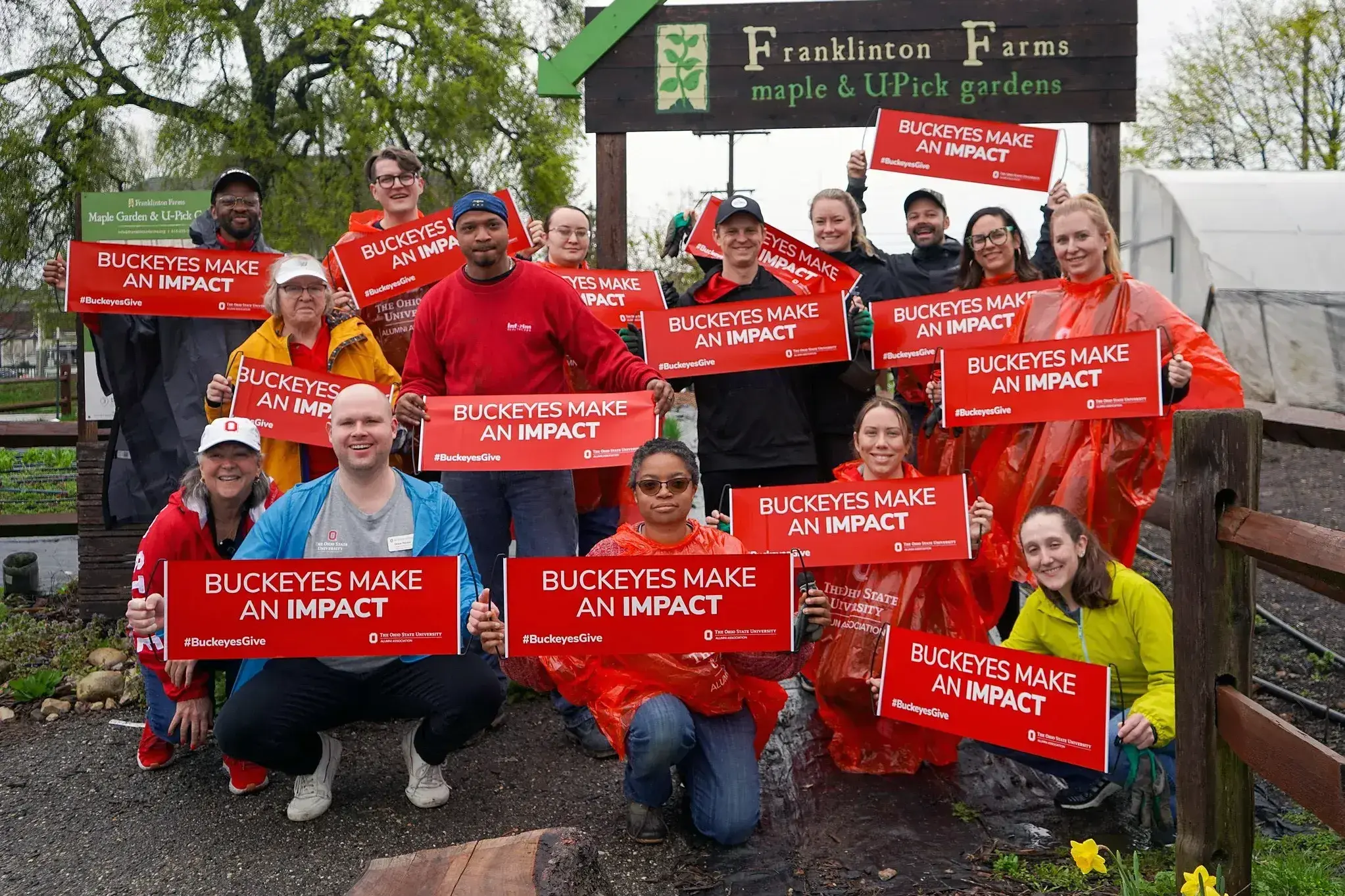 A group of volunteers stands outdoors at Franklinton Farms holding red signs that read “Buckeyes Make an Impact,” with garden beds, trees, and a farm sign in the background.