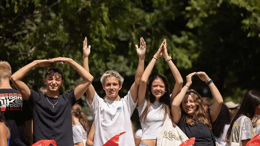 A group of people standing outdoors with arms raised overhead, forming shapes with their hands, with trees in the background.
