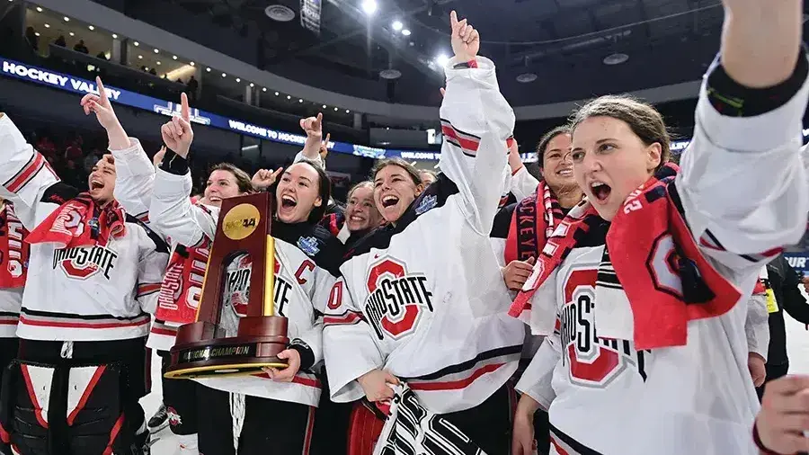 A women’s hockey team wearing Ohio State jerseys celebrates on the ice, raising their arms and holding a championship trophy inside a crowded arena.