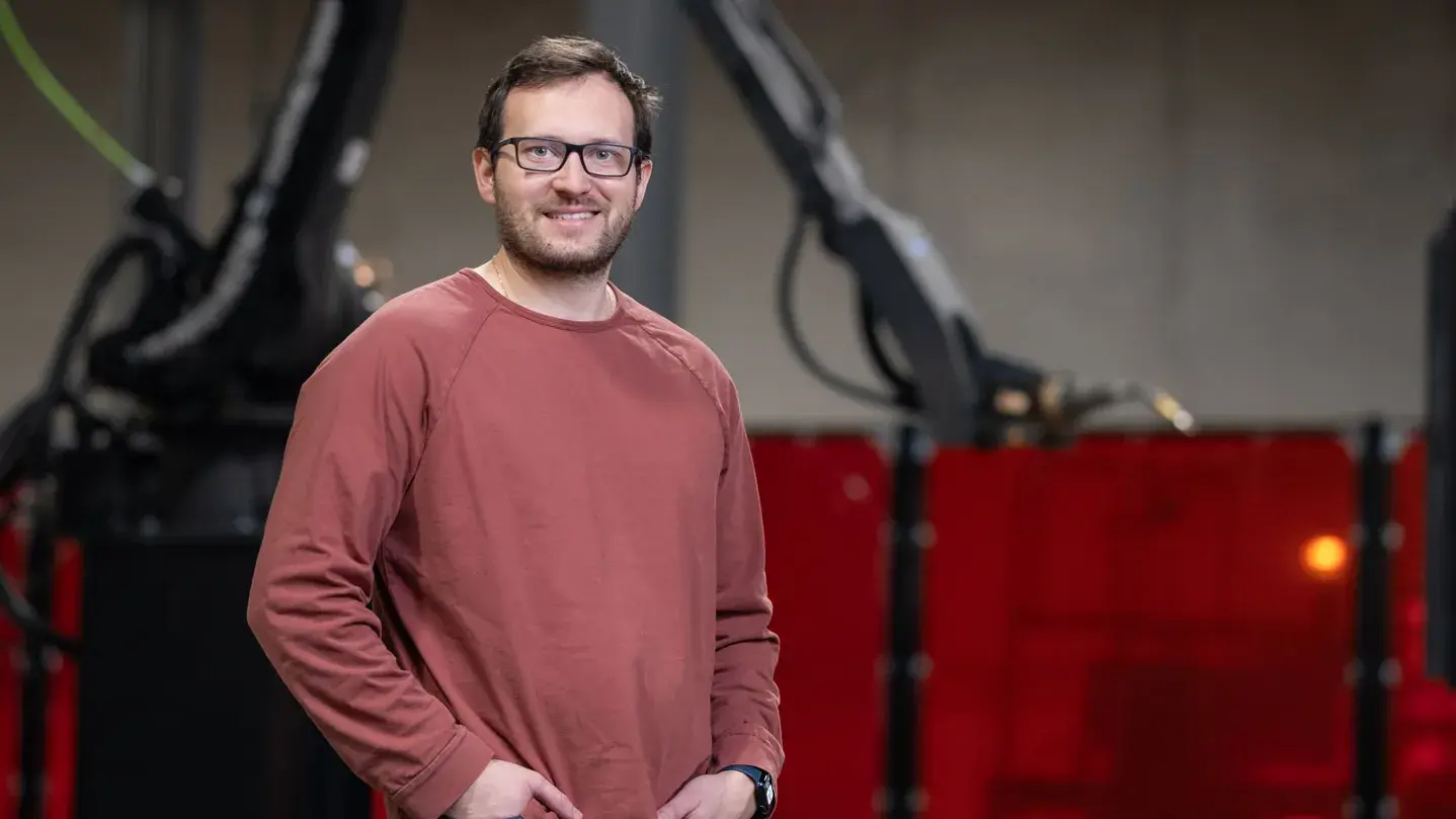 Andy Lonsberry smiles and stands inside a factory with his hands in pockets. Behind him is a giant robotic arm. He wears a sweatshirt, glasses and has neat hair and beard.