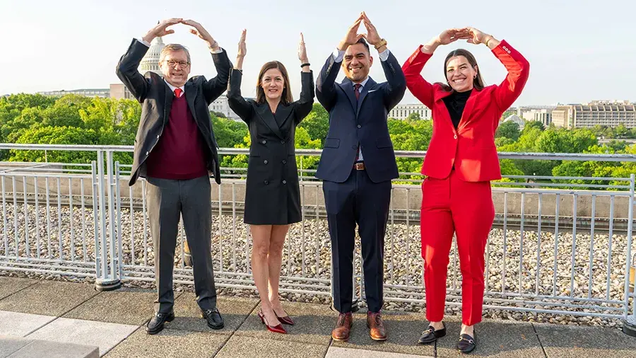 Four people in business clothing stand on an outdoor rooftop terrace, raising their arms overhead in different shapes, with a city skyline and a domed building visible in the background.
