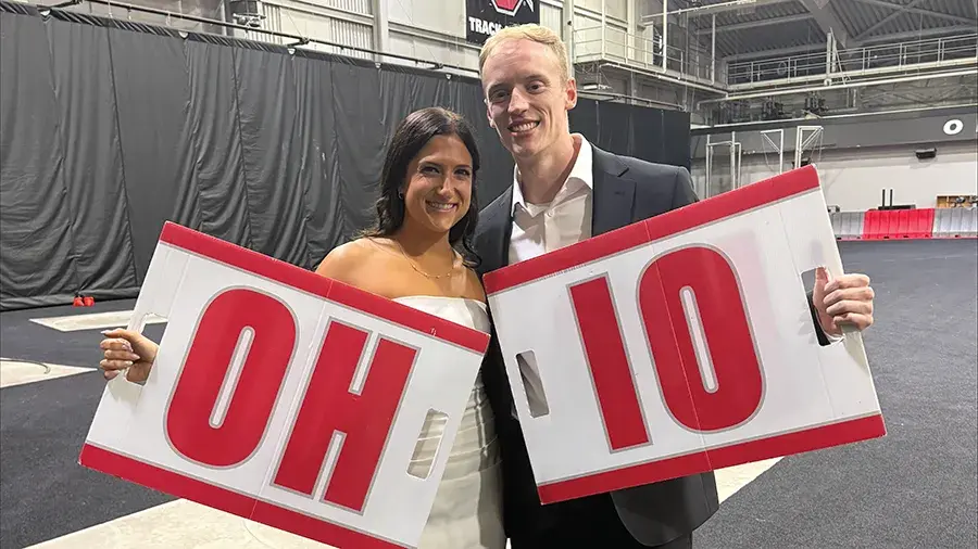 Jordan and Maria stand together in an indoor arena, each holding a large sign that reads “OH” and “IO.” 
