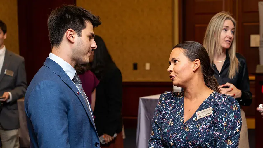 Two adults wearing business attire and name badges talking at an indoor reception or networking event, with other attendees and cocktail tables visible in the background.