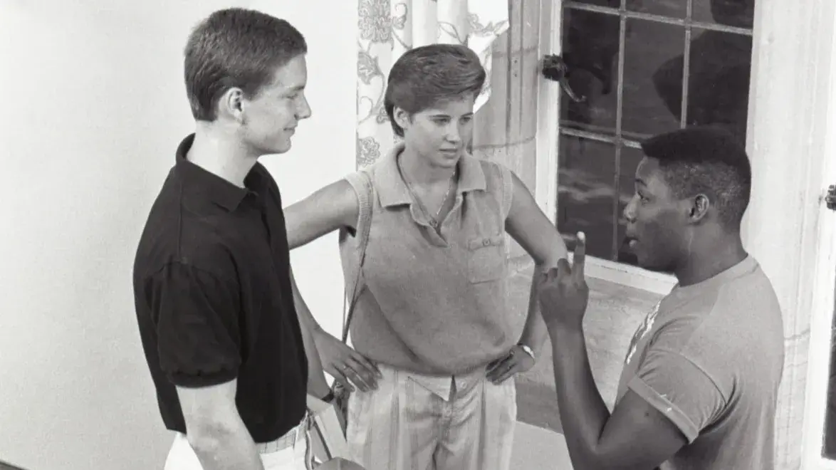 In a photo from the 1980s, three college students engage in conversation on a staircase landing. A large window with patterned curtains is behind them. Of the three, one is woman and two are men; one is Black and two are white. They’re dressed in styles common in the 1980s.