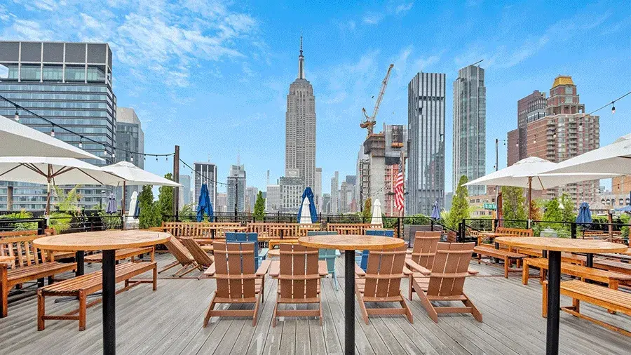 Outdoor rooftop deck with wooden tables and chairs, umbrellas, and lounge seating, overlooking the New York City skyline with the Empire State Building centered in the background.