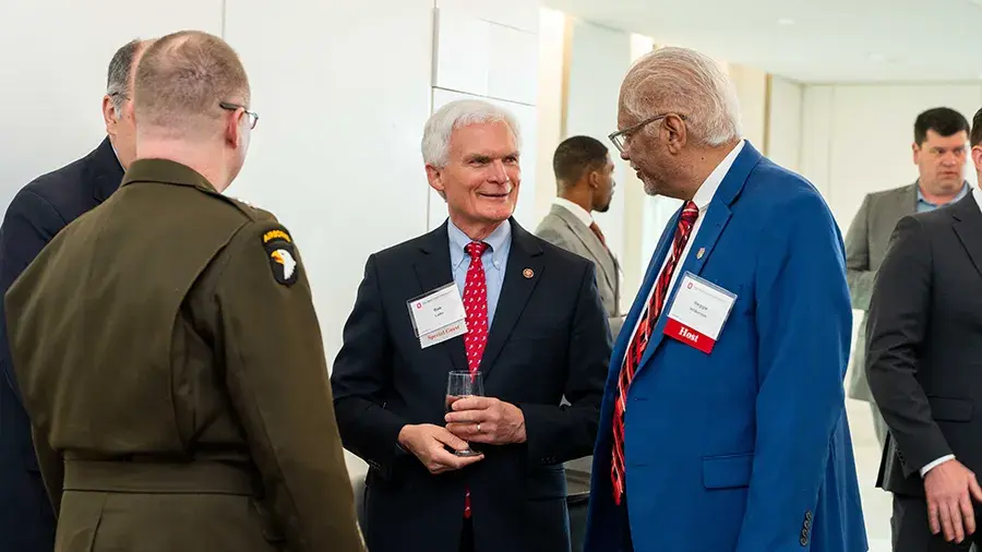 Group of people in formal attire and a military-style uniform standing and talking at an indoor event, wearing name badges with red labels.