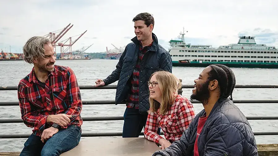 Four people in casual jackets and plaid shirts gathered by a waterfront railing, with a large ferry boat and port cranes visible in the background.