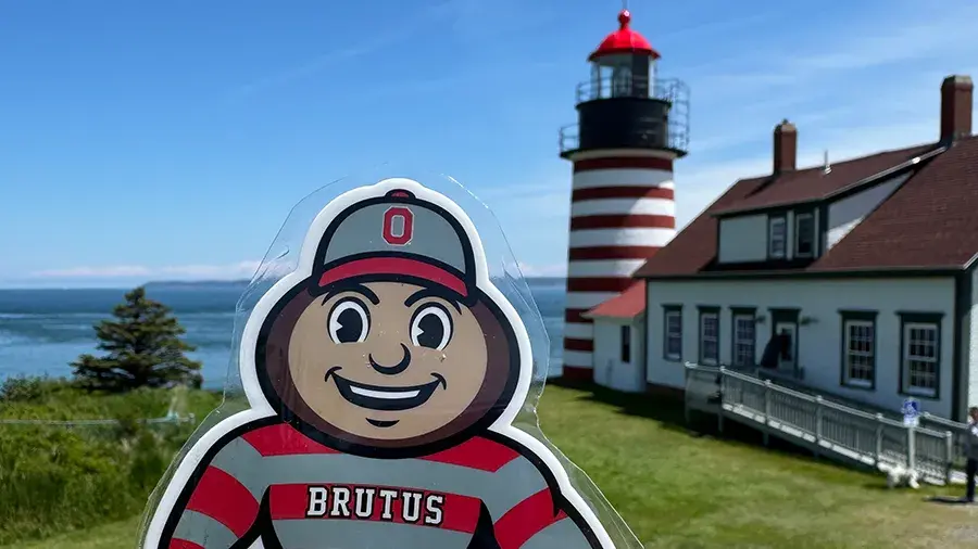 A close-up of a Brutus Buckeye cutout held in front of a coastal lighthouse.