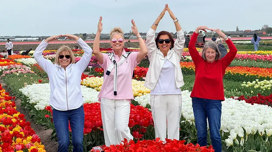 Four people standing in rows of colorful tulips, each raising their arms overhead to form rounded or triangular shapes, with fields of red, yellow, white, and pink flowers stretching into the background under an overcast sky.