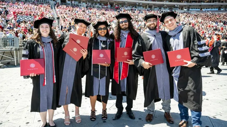 A group of graduates wearing black graduation gowns and caps stand together on a stadium field, each holding a red diploma cover, with a large crowd seated in the stadium behind them.
