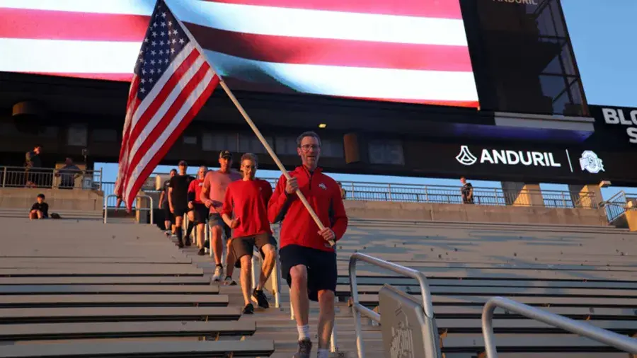 Participant carrying a large American flag leads a group walking down stadium steps at Ohio Stadium.