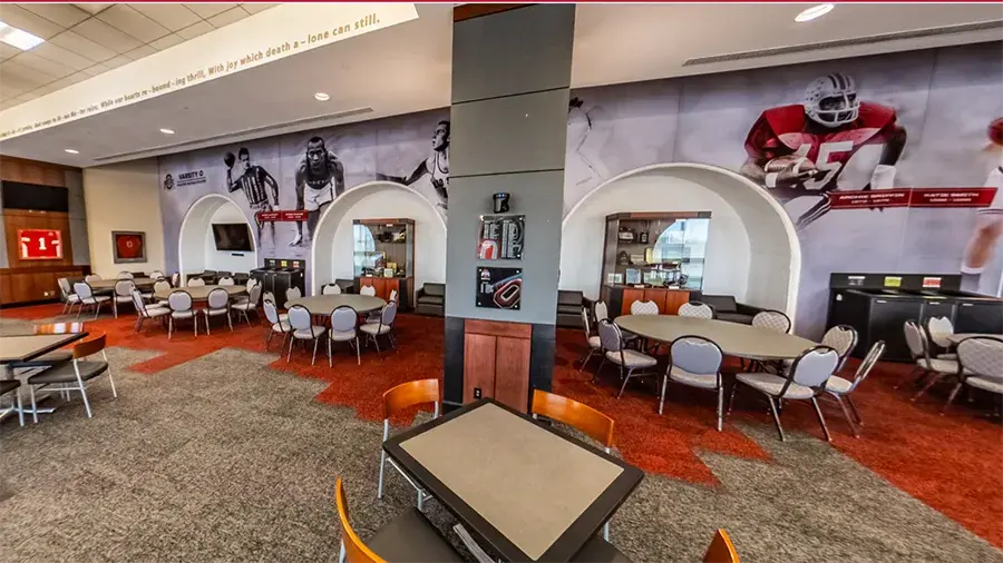 Spacious sports-themed lounge or event room with round and rectangular tables and gray upholstered chairs arranged across a red and gray carpet.