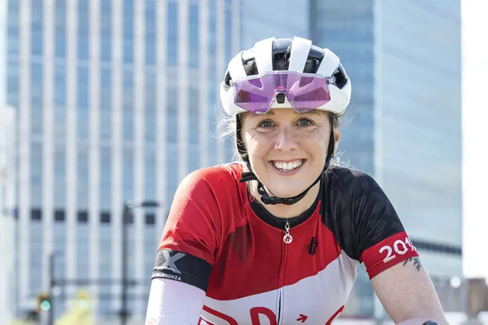 A woman leans on her bicycle handles while wearing a riding helmet and jersey. She smiles as she poses for this photo in front of Ohio State's new hospital tower