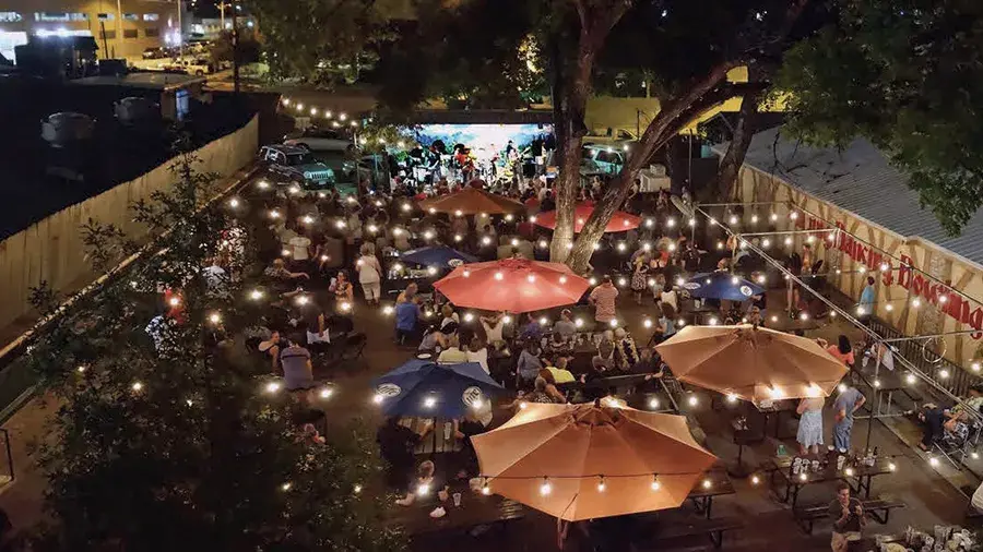 A nighttime outdoor gathering in a courtyard filled with picnic tables, string lights and large patio umbrellas.