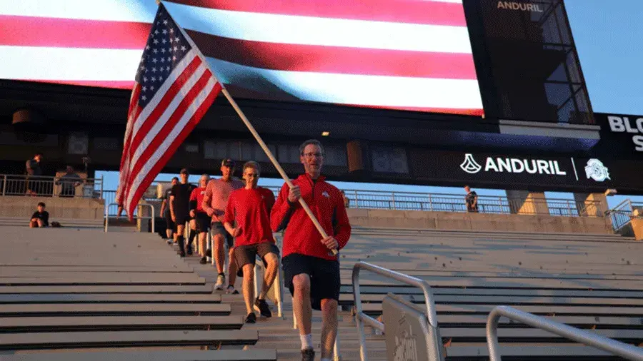 People carrying an American flag descend stadium steps beneath a giant U.S. flag display.