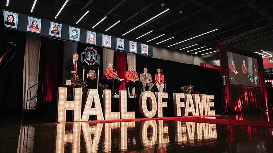 Illuminated “HALL OF FAME” letters stretch across the front of a stage where several speakers sit in a panel discussion beneath Ohio State logos.