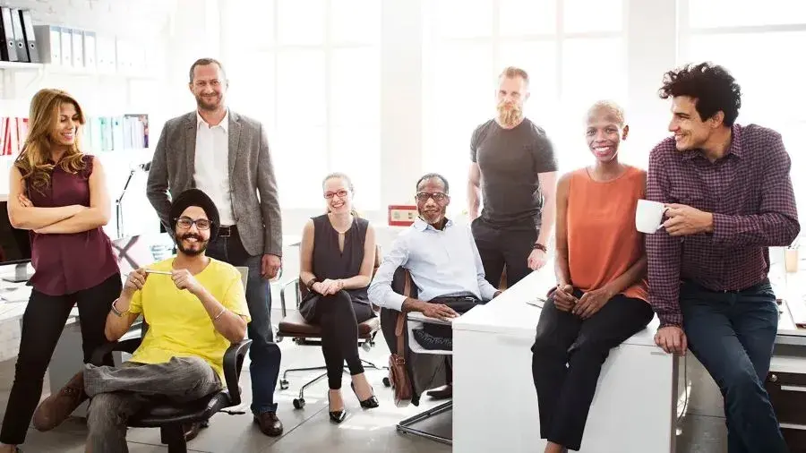 Group of eight coworkers gathered casually in a bright office space, some sitting and some standing around desks, with laptops, chairs, and office supplies visible in the background.